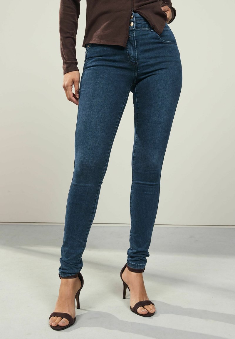 Smala blå denimjeans med hög midja, fem fickor och sömlös design, kombinerade med svarta högklackade sandaler med remmar.