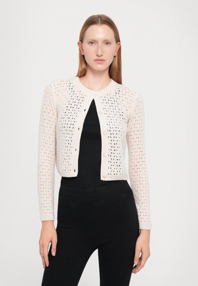 Vaaleanpunainen neulecardigan, jossa on tekstuuri- ja avoimesta työstä koostuva kuvio, pyöreä pääntie ja nappikiinnitys, käytetty mustan paidan ja leggingsien päällä.