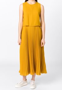 Vestido amarillo sin mangas con falda plisada, hecho de un tejido ligero. Llevado con zapatillas blancas, tiene un escote redondeado.