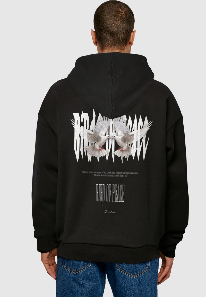 Černá oversized mikina s kapucí, na zadní straně s grafikou dvou bílých holubic, nad nimiž je text a zubatý designový prvek.
