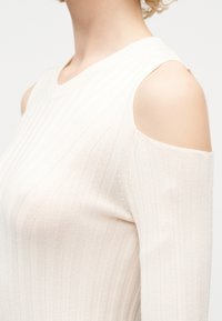 Femme portant un haut en maille côtelée crème avec une découpe épaule dénudée, laissant la peau nue sur le haut du bras et l'épaule.