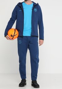 Survêtement bleu marine en tissu doux, comprenant un sweat à capuche zippé et un t-shirt turquoise à manches courtes. Comprend un logo Nike noir et un ballon de football.