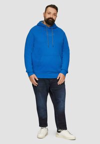 Niebieska bluza z kapturem, posiadająca kieszeń z przodu i sznurki, w połączeniu z ciemnymi dżinsami i białymi tenisówkami. Gładka tekstura, casualowy krój.