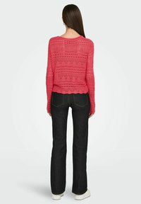 Roze gebreide trui met opengewerkte patronen; gedragen met donkere wijde jeans en witte sneakers. Golvende zoom en losse pasvorm.