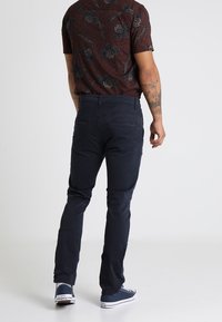 Tummansiniset puuvillahousut, joissa on kapea malli, näkyvät takataskut ja hienot ompeluyksityiskohdat, yhdistettynä kuviolliseen lyhythihaiseen paitaan.