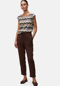Top a maniche corte con un motivo a onde multicolore, abbinato a pantaloni marroni con cintura regolabile. Il modello posa con le mani in tasca.