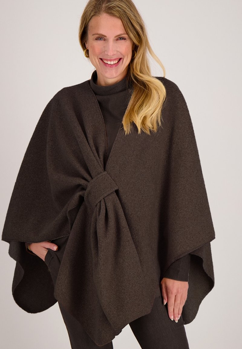 Lachende vrouw met lang blond haar, gekleed in een donkergroene wollen cape, gestrikt aan de voorkant, over een bijpassende top met lange mouwen en broek.