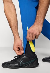 Αθλητικό παντελόνι σε έντονο μπλε με κίτρινη λεπτομέρεια στο αστράγαλο, με κωνικό σχέδιο. Ένα άτομο ρυθμίζει το παντελόνι κοντά σε ένα μαύρο αθλητικό παπούτσι.