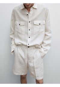 Lichtbeige linnen shirt met een button-down ontwerp, twee borstzakken en zwarte knopen. Gecombineerd met bijpassende shorts in dezelfde stof.