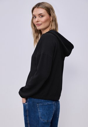 Kobieta o blond włosach, ubrana w czarną bluzę z kapturem i niebieskie dżinsy, stojąca w zrelaksowanej pozie na jasnym, jednolitym tle.