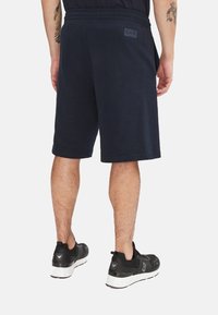 Pantaloni corti di un blu scuro realizzati in tessuto morbido, con una vita elastica e una piccola etichetta con il logo EA7 sul retro. Indossati con scarpe da ginnastica nere.