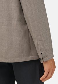 Šedá, texturovaná košile se strukturovaným střihem. Má zapínací manžety a detail bočního švu, v kombinaci s tmavými kalhotami.