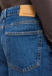 Μπλε τζιν με πίσω τσέπη και δερμάτινη ετικέτα με την ένδειξη "NOISY MAY", φορεμένη με σκούρο καφέ τοπ.