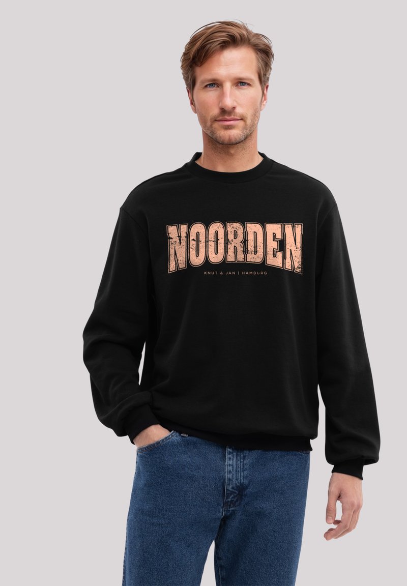 Schwarzer Sweatshirt mit Rundhalsausschnitt, ausgestattet mit einem abgenutzten "NOORDEN"-Grafikdesign in Orange. Kombiniert mit blauen Jeans, die eine lockere Passform zeigen.