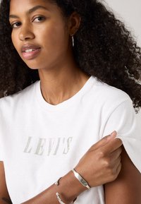 T-shirt blanc avec le logo "LEVI'S" en détails brillants ; le mannequin porte des boucles d'oreilles en argent et un bracelet texturé en argent. Cheveux bouclés visibles.