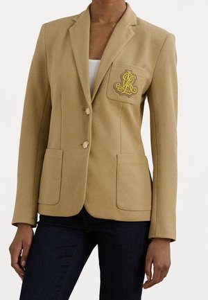 Sandfarvet skræddersyet blazer med to forlommer, guldknapper og broderet gul våbenskjold på brystlommen over hvid top og mørke jeans.