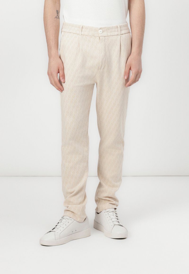Pantaloni beige a fantasia con tessuto testurizzato, dotati di chiusura con bottone e passanti per cintura, abbinati a sneakers bianche.