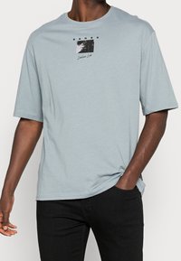 T-shirt à manches courtes bleu clair en matériau doux. Présente un design graphique en noir et blanc à l'avant. Coupe ample, col rond.