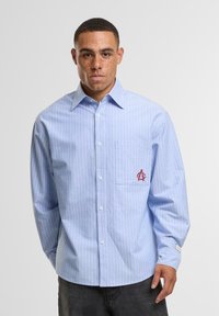 Light blue long-sleeve shirt with white vertical stripes, button-up front, left chest pocket featuring a red embroidered logo.