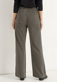 Vrouw in hoge taille wijde broek met een zwart-beige houndstoothpatroon, gecombineerd met zwarte schoenen en een zwart langemouwen top.