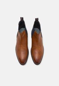Botas de cuero marrón hasta el tobillo con paneles laterales elásticos, punta redondeada y un acento contrastante en azul marino en la parte superior. Textura suave y costuras mínimas.