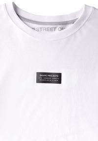 Vit bomull t-shirt med rund halsringning och en liten svart etikett vid bröstet som visar varumärket och designens detaljer.