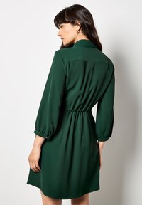 Grünes, knielanges Kleid mit Kragen, elastischem Bund und drei Viertelärmeln. Glatter Stoff mit leichtem Glanz und gerafften Akzenten.