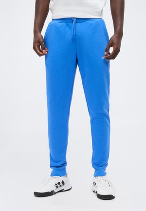 Mann iført lyseblå joggebukser med snøring i livet og hvite sneakers, står mot en ensfarget lys bakgrunn.