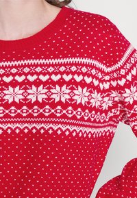 Pull en tricot rouge avec des motifs Fair Isle blancs, comprenant des cœurs et des flocons de neige, avec un col ras du cou et une finition texturée.