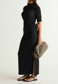 Mustasilkinen maxi-mekko lyhyillä hihoilla ja sivuaukkoilla, yhdistettynä kudottuun ruskeaan clutch-laukkuun ja litteisiin mustiin sandaaleihin.