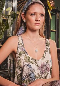 Mlada žena u cvjetnoj haljini, sa odgovarajućom cvjetnom trakom za glavu, naušnicama u obliku cvijeta i ogrlicom, sjedi u zatvorenom prostoru blizu zelenih biljaka i tamnog namještaja.