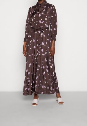 Frau trägt ein langes, dunkelbraunes Kleid mit Knöpfen, verziert mit pinken und weißen Blumenmustern, geschnürter Taille, langen Ärmeln und weißen, offenen Sandaletten mit Absatz.