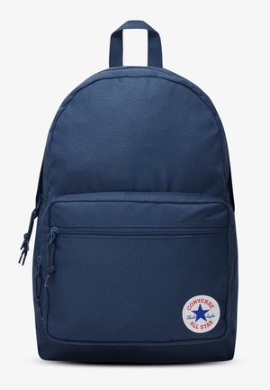 Sac à dos Converse bleu marine avec poche zippée à l'avant et écusson rond du logo Chuck Taylor All Star dans le coin inférieur droit.