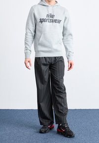 Gray hoodie with "Nike Sportswear" logo, black-and-white striped pants, and black sneakers with red accents. Standing on a blue carpet.