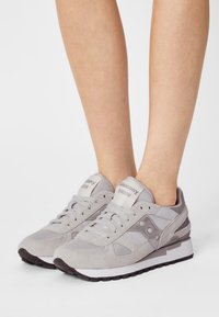 Grijze suede en mesh sneakers met vetersluiting aan de voorkant, gevoerde kraag en zwarte rubberen zool, met "Saucony Shadow" branding op de tong.