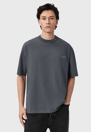 Young man with dark hair wearing a loose dark gray AllSaints t-shirt and black jeans, standing against a plain light background.