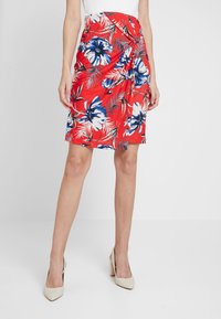 Mujer con falda roja hasta la rodilla con un estampado floral azul y blanco y zapatos de tacón beige, de pie frente a un fondo blanco liso.