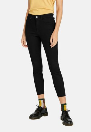 Person trägt schwarze Slim-Fit-Jeans, schwarze Leder-Schnürschuhe und bunte gestreifte Socken, steht vor einem schlichten Hintergrund.