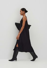 Schwarzes ärmelloses Kleid mit seitlichen Schlitzen, kombiniert mit knielangen schwarzen Stiefeln. Das Model hält eine strukturierte schwarze Tragetasche. Minimalistisches Design.