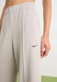 Pantalon de sport gris clair avec une taille élastique, des rayures sur les côtés et un petit logo noir près de la hanche. Texture du tissu lisse et douce.