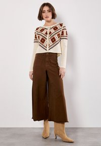 Cremefarbene Häkeljacke mit mehrfarbigen geometrischen Mustern, kombiniert mit hochtaillierten braunen weiten Hosen und beigen Knöchelstiefeln.
