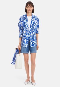 Blazer mit blau-weißem Muster, aus gewebtem Stoff, über einem weißen Oberteil getragen, kombiniert mit Jeansshorts, einer weißen Handtasche und gestreiften Ballerinas.