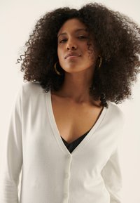 Vit, knäppt cardigan med V-krage, tillverkad av mjukt tyg; har långa ärmar och en figurnära design. Bärs över en svart linne.
