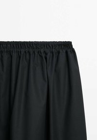 Tissu noir avec une ceinture élastique, froncé en haut, probablement une partie d'une jupe ou d'un pantalon sur fond blanc.