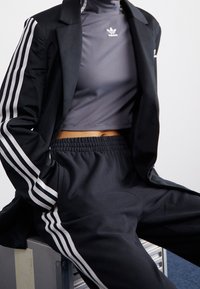 Svart blazer med vita ränder på ärmarna, grå åtsittande polotröja och svarta träningsbyxor med elastisk midja. Slät tyg, sportig design.