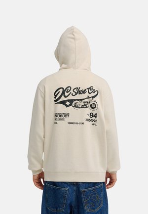 Person trägt einen cremefarbenen Hoodie mit großem schwarzen Motorradmotiv, „DC Shoe Co“-Schriftzug und blauen Jeans mit Designs auf der Gesäßtasche.
