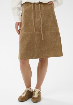 Barna velúr szoknya övvel a derekán, sima textúrával és térd alá érő hosszúsággal. Barna tornacipővel párosítva, harmonizáló megjelenésért.