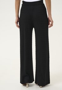 Pantalons larges noirs en tissu texturé, présentant un motif côtelé et une taille élastique. Les pantalons descendent jusqu'au sol avec un design fluide.