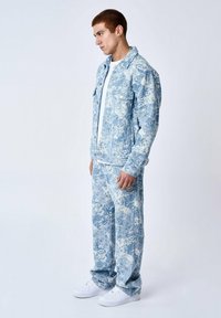 Set di giacca e pantaloni in denim di colore blu chiaro con motivi floreali bianchi. Vestibilità dritta con chiusure a bottoni e tasche laterali. Sneakers bianche indossate.