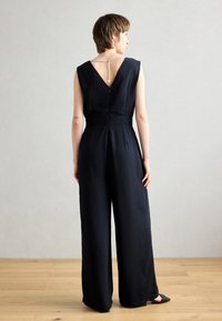 Svart jumpsuit med dyp V-rygg, med kjededetalj. Stoffet er glatt med en avslappet passform og vide ben, som skaper en minimalistisk silhuett.